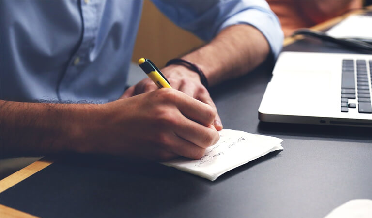 SCORE WP6: A person holding a pen and writing in a notebook on their desk next to their laptop.